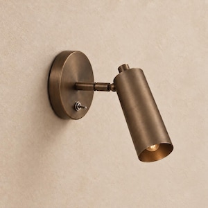 Pode incluir: Um candeeiro de parede com acabamento em latão antigo. O candeeiro possui uma placa de base circular com um pequeno interruptor de alavanca e uma cabeça cilíndrica ajustável fixada por um braço horizontal, direcionando a luz para baixo.
