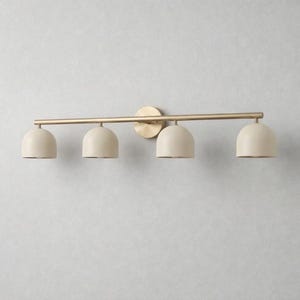 K&ouml;nnte beinhalten: Eine vierflammige Waschtischleuchte mit Messing-Finish. Die Leuchte hat eine horizontale Stange mit vier cremefarbenen, kuppelf&ouml;rmigen Schirmen. Die Leuchte ist auf einer runden Messing-R&uuml;ckplatte montiert.