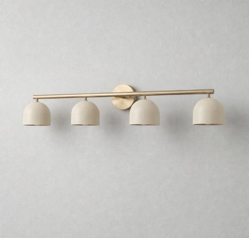 K&ouml;nnte beinhalten: Eine vierflammige Waschtischleuchte mit Messing-Finish. Die Leuchte hat eine horizontale Stange mit vier cremefarbenen, kuppelf&ouml;rmigen Schirmen. Die Leuchte ist auf einer runden Messing-R&uuml;ckplatte montiert.