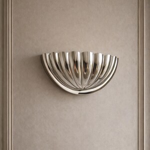 Könnte beinhalten: Eine polierte silberne Wandleuchte mit einem gewellten Design, montiert an einer Wand in neutralen Tönen. Die Leuchte hat eine halbkreisförmige Form mit vertikalen Rippen, die das Licht reflektieren. Die Wand hat ein gerahmtes Paneeldesign.