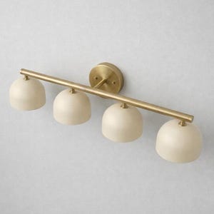 K&ouml;nnte beinhalten: Eine vierflammige Waschtischleuchte mit Messing-Finish. Die Leuchte hat eine horizontale Stange mit vier cremefarbenen, kuppelf&ouml;rmigen Schirmen. Sie ist auf einer runden Messing-R&uuml;ckplatte montiert.