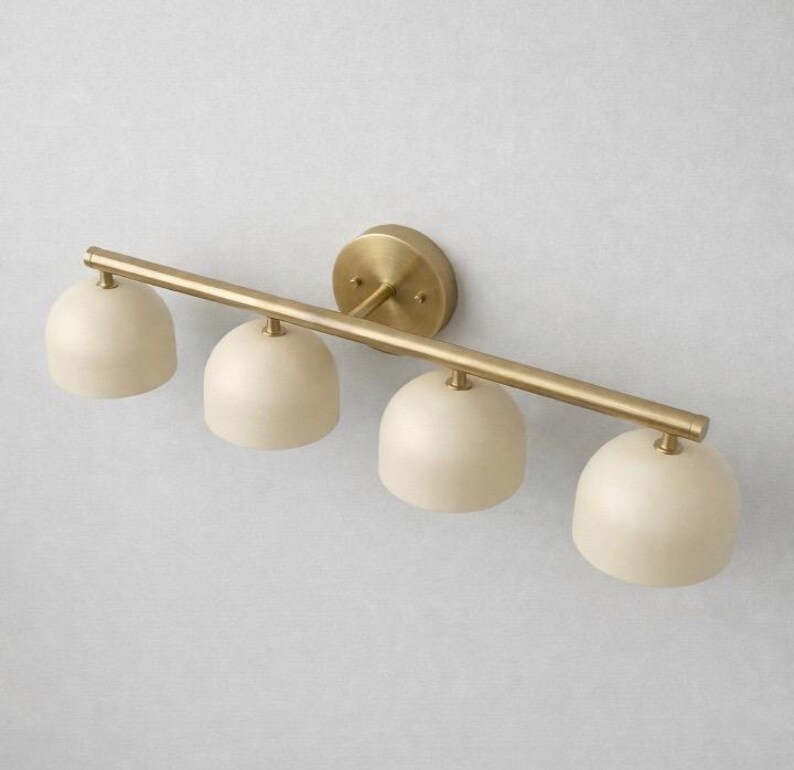 K&ouml;nnte beinhalten: Eine vierflammige Waschtischleuchte mit Messing-Finish. Die Leuchte hat eine horizontale Stange mit vier cremefarbenen, kuppelf&ouml;rmigen Schirmen. Sie ist auf einer runden Messing-R&uuml;ckplatte montiert.