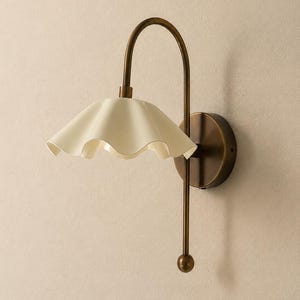 May include: A wall-mounted lamp featuring a curved, antique brass-toned metal arm and a circular wall plate. The light fixture includes a wide, off-white, scalloped-edge shade that flares outward, suspended from the top of the arched metal frame.