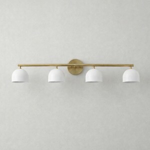 K&ouml;nnte beinhalten: Eine vierflammige Waschtischleuchte mit Messing-Finish. Die Leuchte hat eine horizontale Stange mit vier wei&szlig;en, kuppelf&ouml;rmigen Schirmen. Die Schirme sind gleichm&auml;&szlig;ig &uuml;ber die Stange verteilt. Die Leuchte ist auf einer runden Messing-R&uuml;ckplatte montiert.