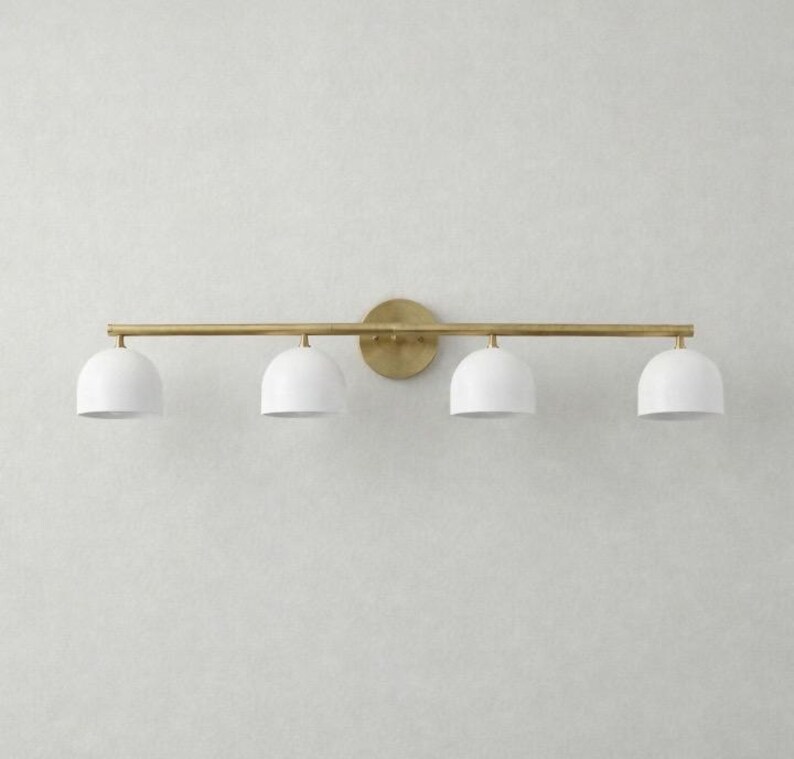 K&ouml;nnte beinhalten: Eine vierflammige Waschtischleuchte mit Messing-Finish. Die Leuchte hat eine horizontale Stange mit vier wei&szlig;en, kuppelf&ouml;rmigen Schirmen. Die Schirme sind gleichm&auml;&szlig;ig &uuml;ber die Stange verteilt. Die Leuchte ist auf einer runden Messing-R&uuml;ckplatte montiert.