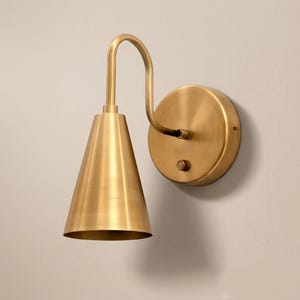 May include: A wall-mounted lamp featuring a brushed brass finish. The fixture consists of a circular wall plate with a small circular switch, connected to a curved arm that supports a downward-facing conical shade in a matching brass tone.