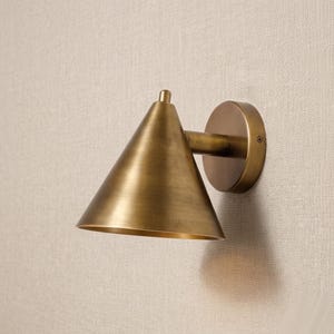May include: A wall-mounted lamp featuring a conical shade made of brushed brass. The fixture includes a circular base plate that attaches to the wall, connected by a short horizontal arm to the angled, downward-facing cone-shaped light housing.