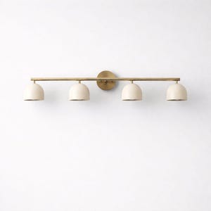 May include: A modern, four-light vanity fixture. The light features a brass-colored horizontal bar and circular wall mount. Four off-white, dome-shaped shades are evenly spaced along the bar. The fixture is designed for bathroom or interior lighting.