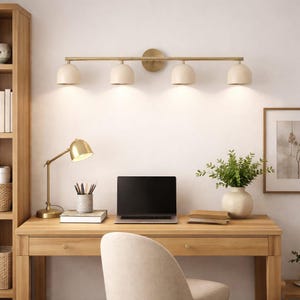 K&ouml;nnte beinhalten: Ein Home-Office-Setup mit einem Holzschreibtisch, einem Laptop, einer Schreibtischlampe und einer Vier-Licht-Waschtischleuchte. Die Waschtischleuchte hat eine Messingoberfl&auml;che und cremefarbene Schirme. Ein gerahmtes Bild und eine Vase mit Gr&uuml;nzeug vervollst&auml;ndigen die Szene.