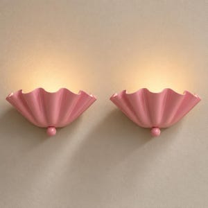 Puede incluir: Un par de apliques de pared con un diseño semicircular en forma de concha con bordes ondulados en un acabado rosa claro sólido. Cada lámpara incluye un pequeño acento esférico en la base y emite un brillo cálido desde la apertura superior.