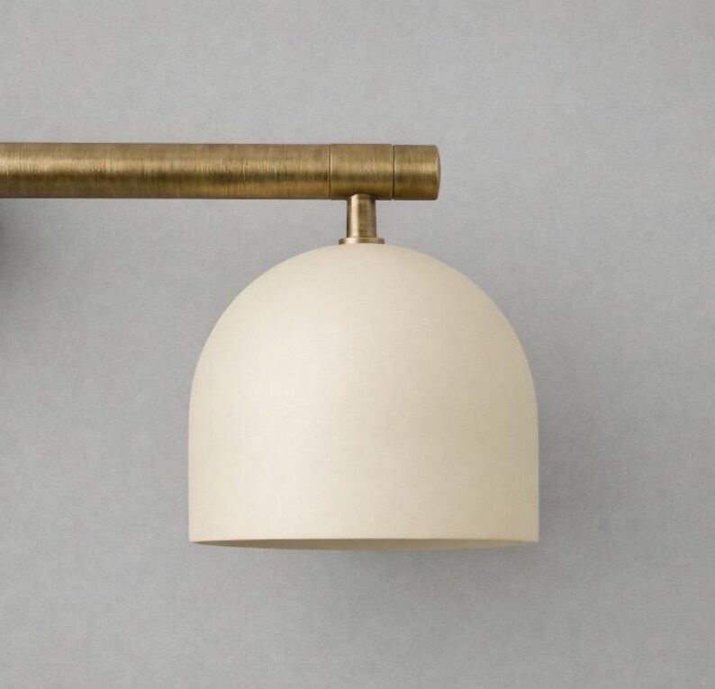 K&ouml;nnte beinhalten: Eine moderne Wandleuchte mit einem cremefarbenen, kuppelf&ouml;rmigen Schirm und einer horizontalen Stange in Messingfarbe. Die Leuchte ist an einer grauen Wand montiert und zeigt ein minimalistisches Design. Die Leuchte ist ideal, um Umgebungslicht zu erzeugen.