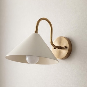 Könnte beinhalten: Wandleuchte mit weißem, konischem Schirm und gebogenem Messingarm. Sie hat einen runden Messingfuß und eine weiße Glühbirne. Das Design ist minimalistisch und modern, passend für verschiedene Innenräume.