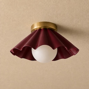 May include: A flush mount ceiling light featuring a deep burgundy, pleated, scalloped metal shade. The fixture has a brushed brass circular base and a central, spherical, frosted white glass bulb that emits a soft, diffused light.