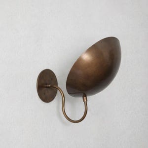 Könnte beinhalten: Bronze Wandleuchte mit geschwungenem Arm und schalenförmigem Schirm. Die Leuchte hat einen kreisförmigen Sockel und eine warme, metallische Oberfläche. Der Schirm ist abgewinkelt und lenkt das Licht nach unten.