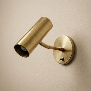 May include: A wall-mounted spotlight fitting with a brushed brass finish. The lamp features a cylindrical head attached to a circular base plate by an adjustable arm. A small metal toggle switch is located on the front of the circular base plate.