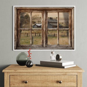 Könnte beinhalten: Ein gerahmter Kunstdruck mit einem rustikalen Holzfenster. Das Fenster zeigt eine ländliche Landschaft mit Scheunen, Zäunen und Feldern. Eine Holzkommode trägt dekorative Gegenstände: Vasen, Bücher, eine Uhr und einen Diffusor.