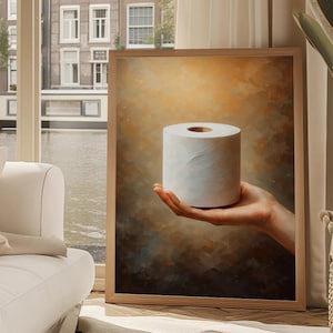 Könnte beinhalten: Ein gerahmter Kunstdruck mit einer Rolle weißem Toilettenpapier, die von einer Hand gehalten wird. Der Hintergrund hat ein warmes, strukturiertes, abstraktes Design mit Braun- und Goldtönen. Das Kunstwerk wird in einem Raum mit Fenster ausgestellt.