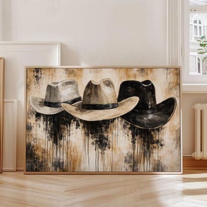 Puede incluir: Una obra de arte enmarcada con tres sombreros de vaquero en tonos grises, beige y negro. Los sombreros están dispuestos horizontalmente sobre un fondo texturizado con vetas verticales marrones y negras. La obra está en un marco dorado.