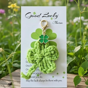 May include: A green crocheted four-leaf clover charm with a gold-colored clasp, attached to a card that reads "Good Lucky Charm". The charm is displayed on a wooden surface with a natural background of flowers and greenery.
