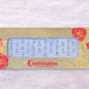 Puede incluir: Una caja rectangular de Combination Designer Bindi. La caja tiene un diseño floral dorado con rosas rojas. En el interior, hay múltiples bindis decorativos plateados en forma de lágrima con un aspecto brillante.