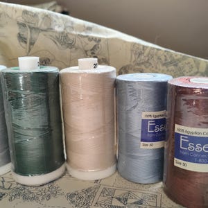 Puede incluir: Una colección de carretes de hilo en varios colores, incluyendo plata, verde, beige, azul y marrón. Los carretes están etiquetados con información del producto, incluyendo el tipo y tamaño del hilo. El hilo está envuelto en plástico transparente.