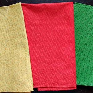 May include: Three folded fabric cloths in yellow, red, and green. The yellow cloth has a dotted pattern, while the red and green cloths have a textured weave. The cloths are likely kitchen towels or napkins.