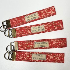 May include: Four red fabric keychains with gold polka dots and bronze hardware. Each keychain has a different inspirational quote: "NO GUTS NO STORY", "YOU ARE DOING GREAT", "AND STILL, I RISE", and "TAKE THE RISK".