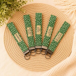 May include: Five fabric keychains in a green and white dashed pattern, each featuring a rectangular tan label with black text. The labels read: FLOURISH, RELAX SLOW DOWN, DREAM BIG, YOU ARE YOUR BEST THING, and BE A VOICE NOT AN ECHO. Each has a metal ring.
