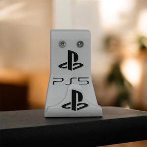 Puede incluir: Un soporte blanco para consola PS5 con el logotipo de PlayStation y el texto "PS5" en negro. El soporte tiene dos tornillos en la parte superior y una base curva. Está diseñado para sostener una consola PS5.