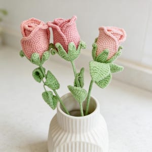 Puede incluir: Tres rosas rosas de ganchillo en un jarrón blanco. Las flores tienen tallos y hojas verdes. Las rosas hechas a mano son un elemento decorativo encantador.