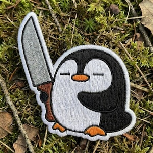 May include: Embroidered patch featuring a cartoon penguin with a large knife. The penguin has a black and white body, orange feet, and a closed-eye expression. The knife has a silver blade and a brown handle. The patch is set against a background of green moss and leaves.