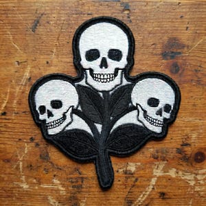 May include: Embroidered patch featuring three white skulls with black eye sockets and teeth, arranged around a central stem with black leaves. The patch has a black border and is set against a wooden background.