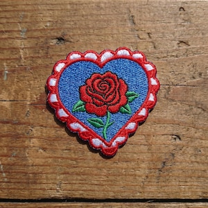 May include: Embroidered patch in the shape of a heart with a red rose in the center. The heart has a blue background and a scalloped red border with white accents. The rose has green leaves.