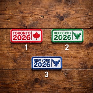 May include: Three rectangular embroidered patches with white borders. The top patch is red with "TORONTO 2026" and a maple leaf. The middle patch is green with "MEXICO CITY 2026" and an eagle. The bottom patch is blue with "NEW YORK 2026" and an eagle.