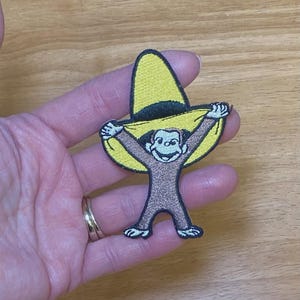 May include: Embroidered patch featuring a cartoon monkey wearing a yellow sombrero. The monkey is brown with a smiling face and is holding the hat up with both hands. The patch has a black border and is on a light brown background.