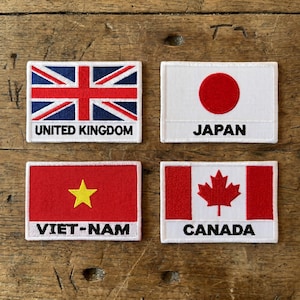 May include: Four embroidered patches featuring national flags. The flags include the United Kingdom, Japan, Vietnam, and Canada. Each patch has a white border and the name of the country is printed in black text below the flag.