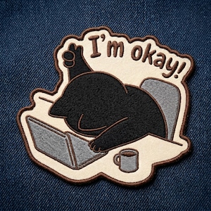 May include: Embroidered patch featuring a black cartoon character slumped over a laptop with the text "I'm okay!". The patch includes a coffee mug and a desk, all outlined in brown.