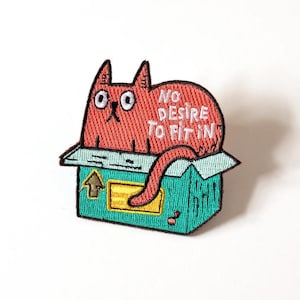 May include: Embroidered patch featuring a red cat peeking out of a teal box. The cat has large eyes and the text "NO DESIRE TO FIT IN" is embroidered on the cat's body. The box has an arrow and a yellow rectangle.