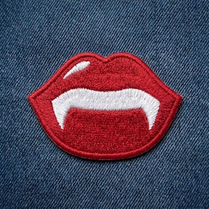 May include: Embroidered patch featuring a pair of red lips with white fangs. The patch is outlined in red and is set against a blue denim background. This patch is suitable for jackets, bags, or other fabric items.