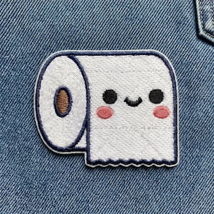 May include: Embroidered patch featuring a smiling roll of toilet paper with black eyes, a brown center, and pink cheeks. The patch has a white background with a navy blue border and is set against a blue denim background.