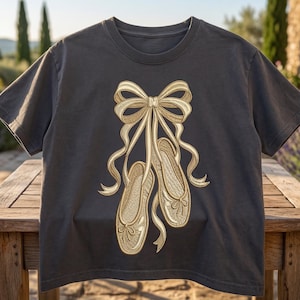 May include: Dark gray t-shirt featuring a gold embroidered design of ballet shoes tied with a bow. The shirt is displayed on a wooden table outdoors, with a blurred background of a stone building and lavender bushes.