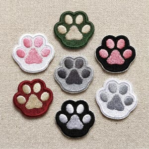 May include: A collection of seven embroidered paw print patches in various colors. The patches feature a paw print design with a white border and are in shades of pink, green, gray, red, and black. These patches are ideal for adding to clothing or accessories.