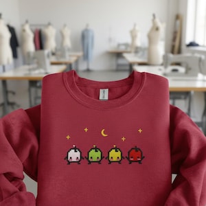 May include: A maroon sweatshirt featuring a pixelated design of four colorful characters with a crescent moon and stars. The characters are in pink, green, yellow, and red. The sweatshirt is displayed in a studio setting.
