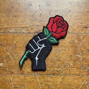 May include: Embroidered patch featuring a black fist holding a red rose with green leaves and stem. The patch is on a wooden surface. The fist is outlined in white, and the rose is detailed with black stitching.