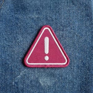 May include: A pink embroidered patch in the shape of a triangle with a white exclamation point inside. The patch has a white border and is set against a blue denim background.