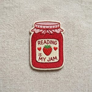 May include: Embroidered patch featuring a red jar of jam with a pink lid and the text "READING IS MY JAM." The patch includes a strawberry and two hearts. The patch is on a beige fabric background.