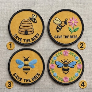 May include: Four round embroidered patches with a yellow background and black borders. Each patch features a bee-related design and the text "SAVE THE BEES". Designs include a beehive, a bee with a flower, and bees with blue wings.