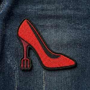 May include: Embroidered patch of a red high-heeled shoe with a black outline and a trident heel. The patch is set against a blue denim background, showcasing the detailed stitching and design of the shoe.