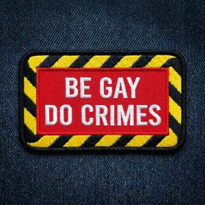 May include: Embroidered patch with the text "BE GAY DO CRIMES" in white on a red background. The patch has a black border with yellow and black diagonal stripes. The patch is rectangular with rounded corners.