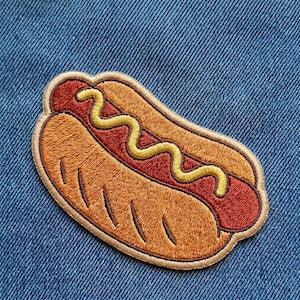 May include: Embroidered patch featuring a hot dog with a red sausage, yellow mustard, and a tan bun. The patch is outlined in a darker tan color and is set against a blue denim background.